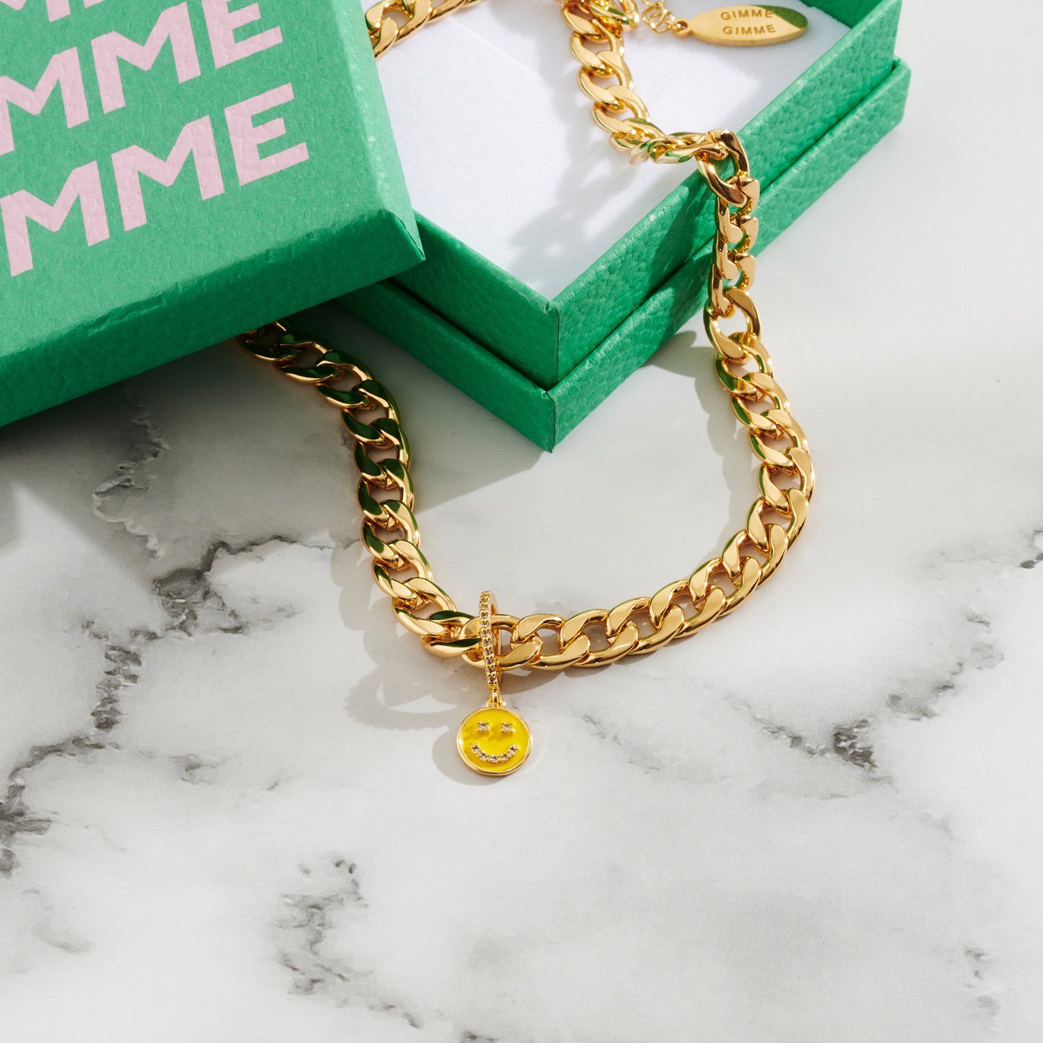 Lifestyle image of chunky gold chain necklace sitting in a green box with smiley face charm.