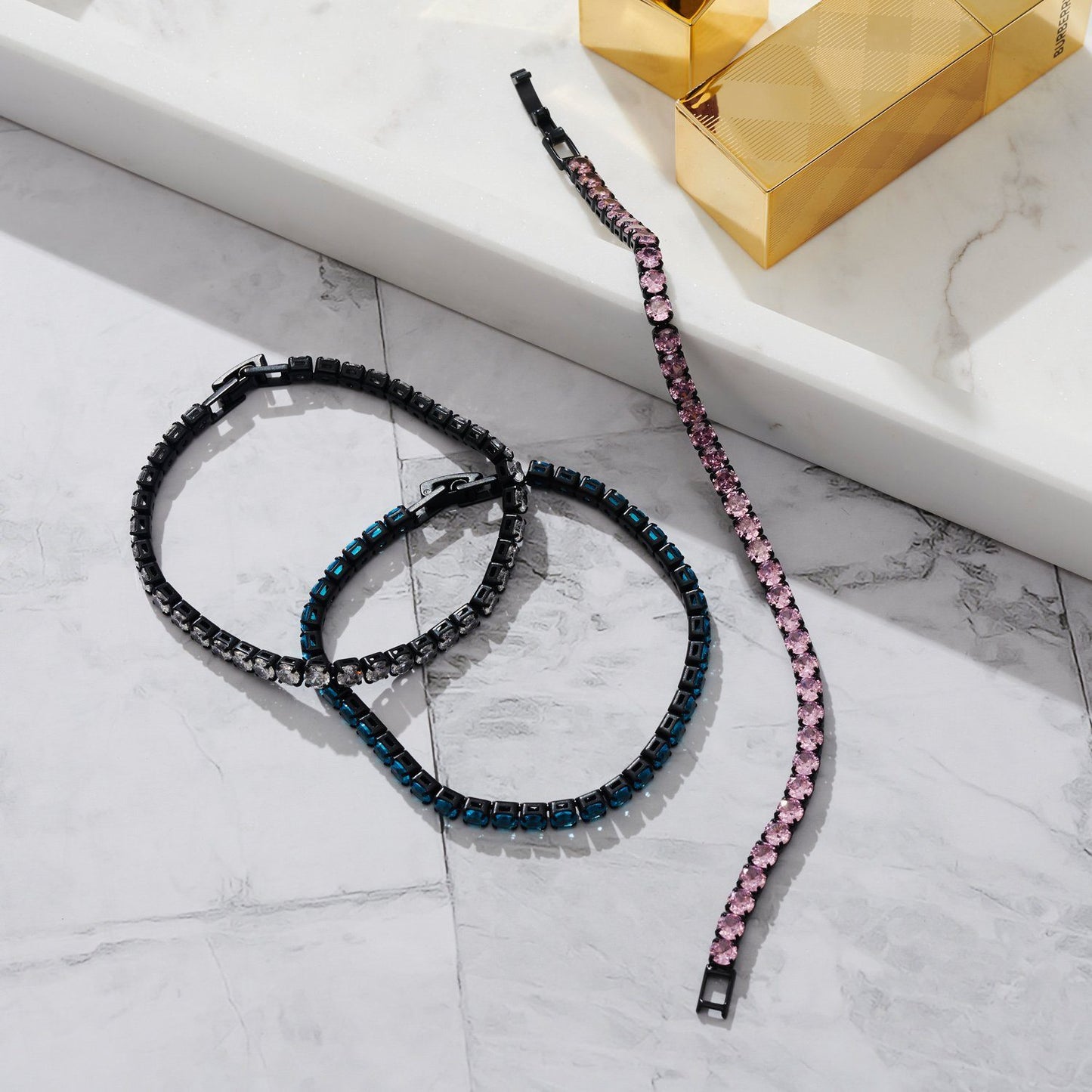 Lifestyle image of three tennis bracelets with black hardware laying on marble bench top.
